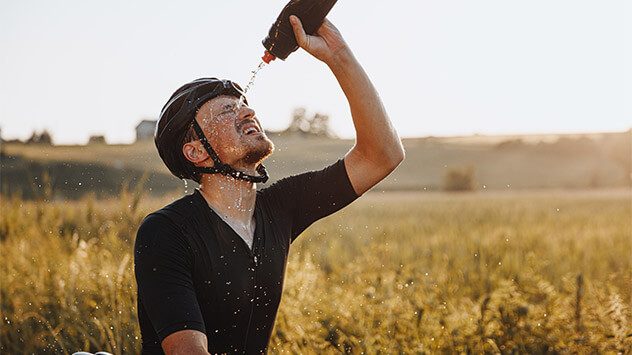 Radfahrer mit Helm kühlt sich in der Sommerhitze mit Wasser aus seiner Trinkflasche ab. (c) LovetheLifeyouLive/Shutterstock.com