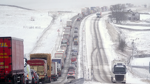 Long line of cars and lorries stuck on a snow-covered motorway, with icy lanes, reduced visibility, and snow-covered fields and hills on both sides.