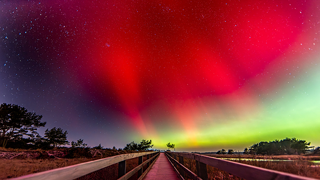 Træbro om natten under røde og grønne nordlys. Stjernehimmel over et åbent landskab.