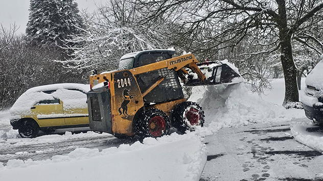 Radlader räumt Schnee von einer verschneiten Straße. Autos und Bäume sind stark mit Schnee bedeckt. Große Schneemengen nach anhaltendem Schneefall.