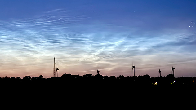 Leuchtende Nachtwolken über Fehmarn mit Windrädern als Silhouetten am Horizont.