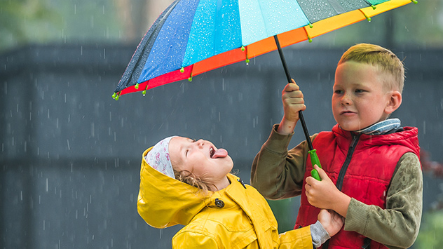 Kinder unter buntem Regenschirm, eines fängt Regentropfen mit der Zunge auf.