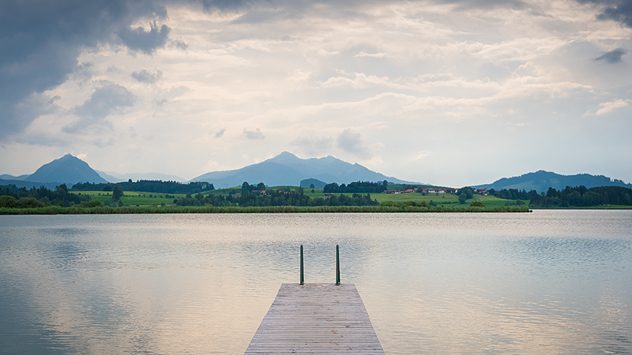 Holzsteg ragt in einen ruhigen See mit Blick auf grüne Hügel und Berge unter bewölktem Himmel. (c) Traveller Martin/Shutterstock.com