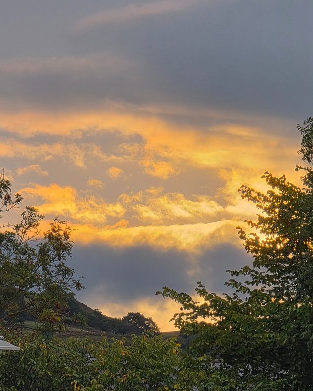 Golden sunset light illuminates layered clouds above green trees and distant hills, with darker gray cloud bands overhead.