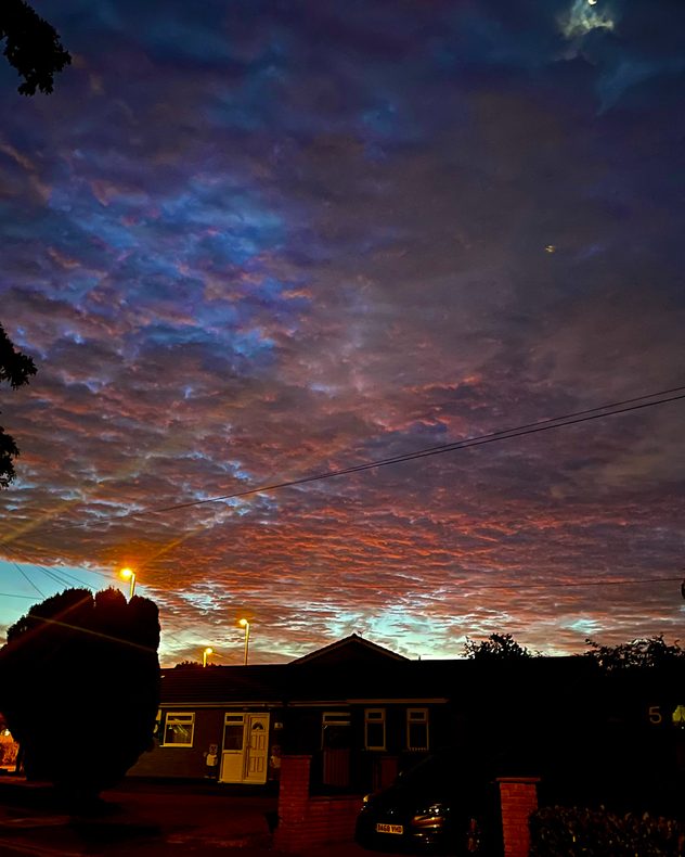 Deep purple, blue and red twilight clouds spread above silhouetted homes, glowing streetlights and power lines, with a crescent Moon visible overhead.