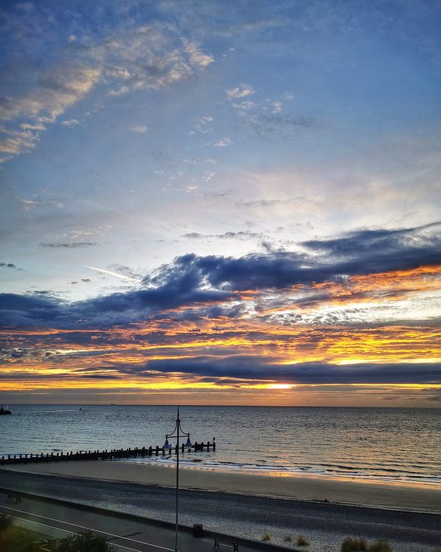 Golden-orange sunset beneath layered blue-gray clouds over a calm sea, sandy beach and wooden groynes along the shoreline.
