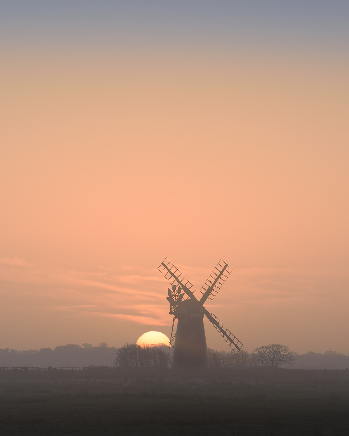 Silhouette of a traditional windmill at sunset, with the sun low on the horizon behind it, orange and peach sky fading into mist, and dark trees and fields in the foreground.