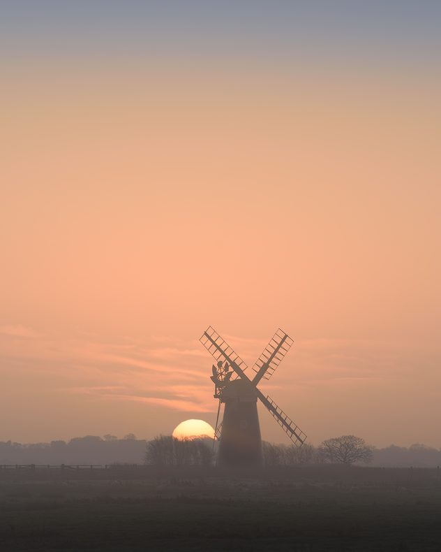 Silhouette of a traditional windmill at sunset, with the sun low on the horizon behind it, orange and peach sky fading into mist, and dark trees and fields in the foreground.