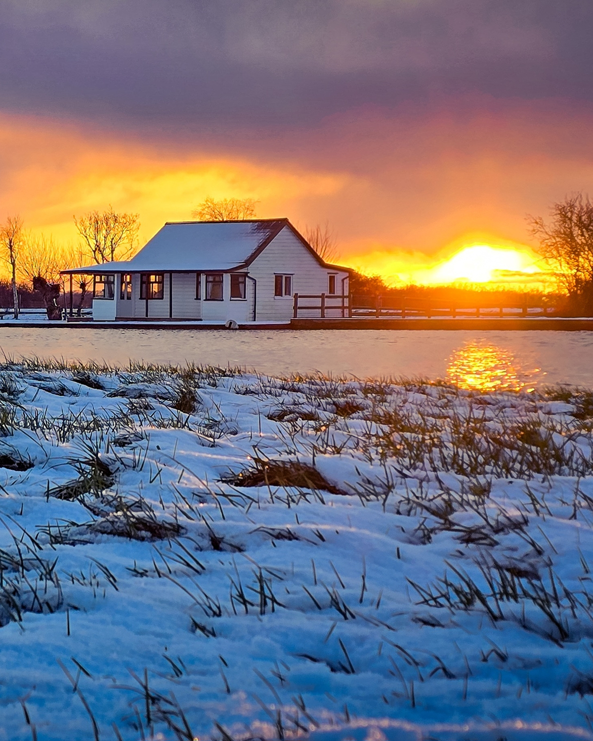 Title: snowy cottage sunset Alt text: Small white cottage beside a frozen pond at sunset, with snow-covered grass in the foreground and vivid orange and yellow sky glowing behind dark winter trees.