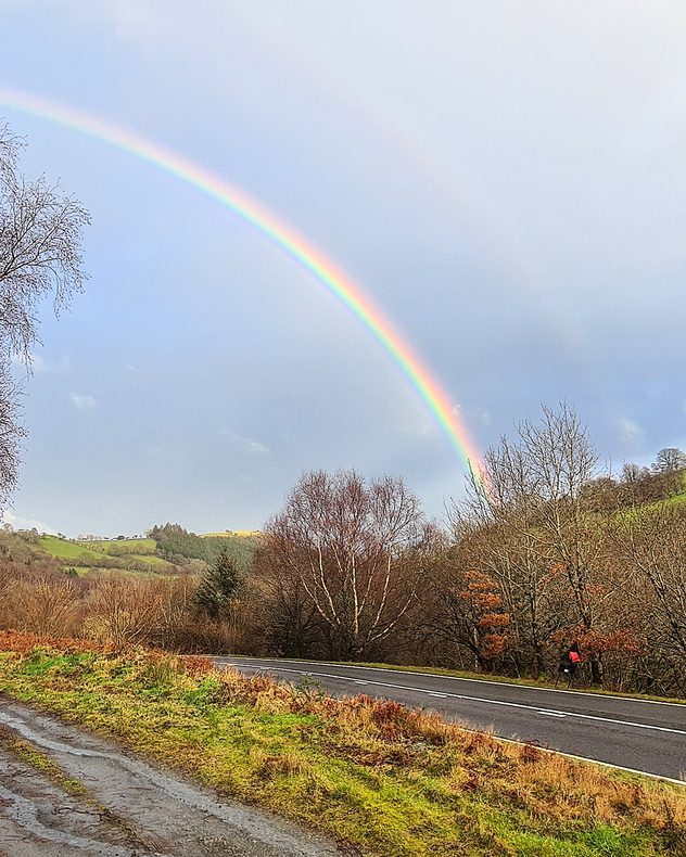 Bright rainbow arching across a grey sky above rolling green hills, bare trees, and a winding country road in the foreground.