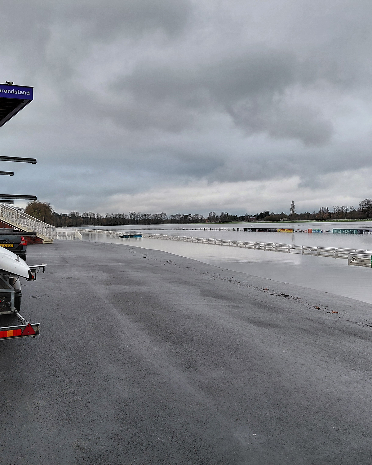 Floodwater covering a racecourse and track rails under a grey overcast sky, with grandstand seating and parked vehicles on higher ground in the foreground.