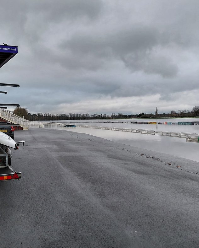 Floodwater covering a racecourse and track rails under a grey overcast sky, with grandstand seating and parked vehicles on higher ground in the foreground.