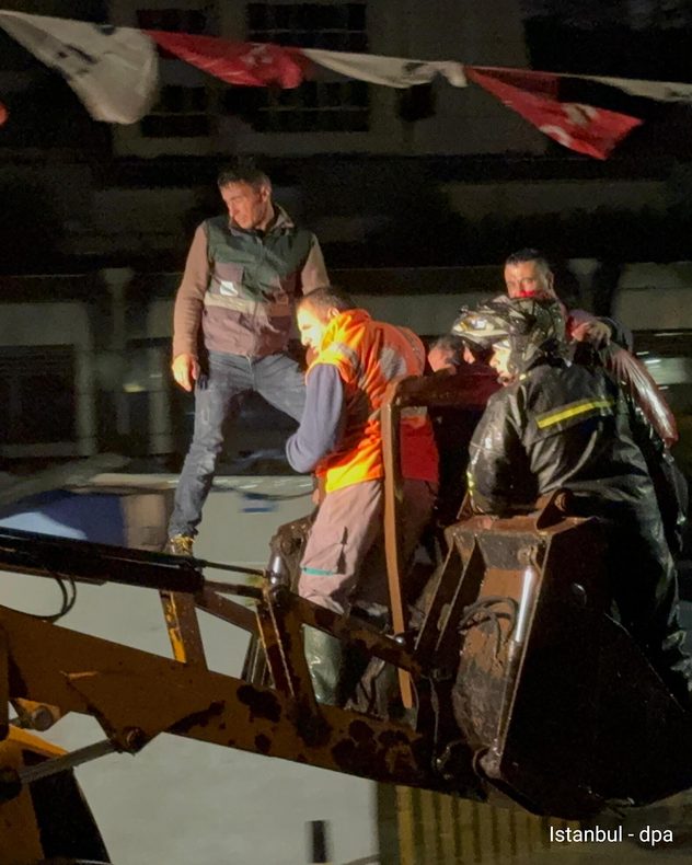 Varios hombres, entre ellos miembros de los servicios de emergencia con trajes de protección, se encuentran de noche sobre la pala de una cargadora sobre ruedas en Estambul. Están situados por encima de una calle inundada.