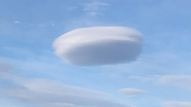 Nube lenticular suave, con forma de lente, que se cierne sobre un cielo azul, con una forma ovalada aplanada distintiva y bordes suaves en capas, que destaca entre las nubes más finas que la rodean.