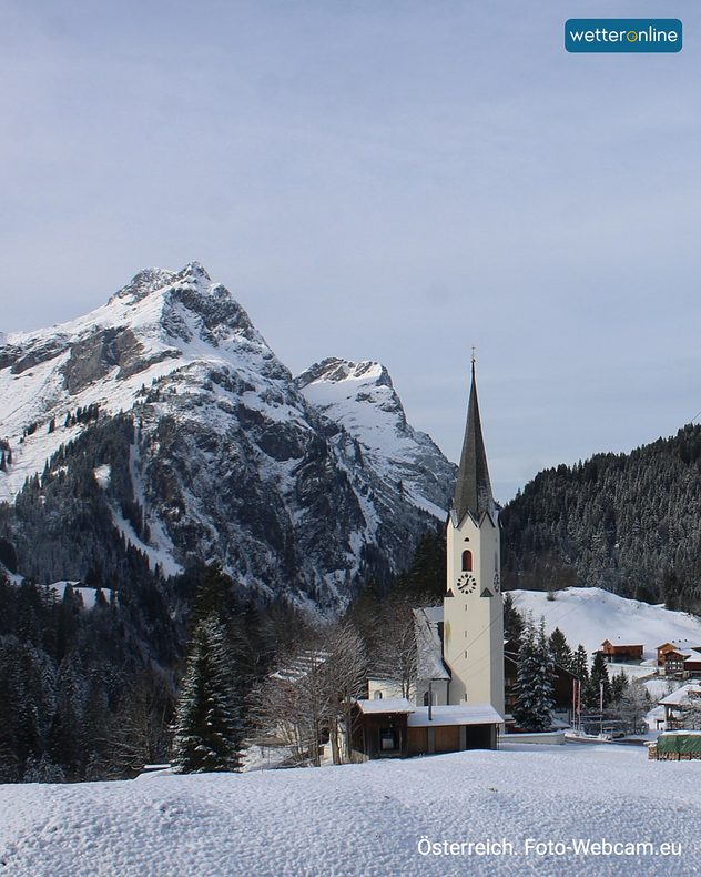 erschneite Kirche in einem österreichischen Alpenort mit hohen Bergen im Hintergrund.