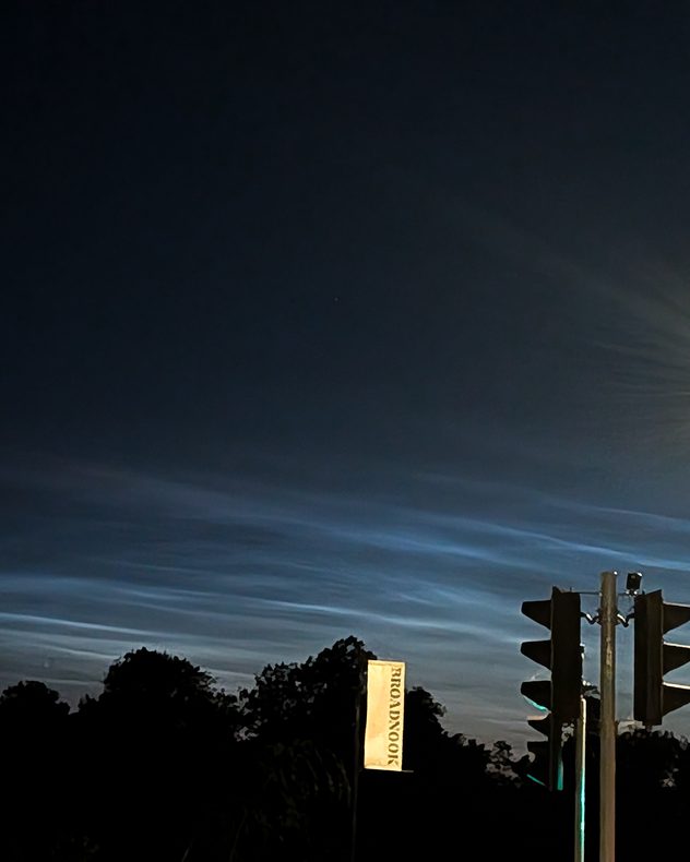 Electric-blue noctilucent clouds streching across a dark twilight sky above silhouetted trees and traffic lights, with delicate rippled bands glowing near the horizon.