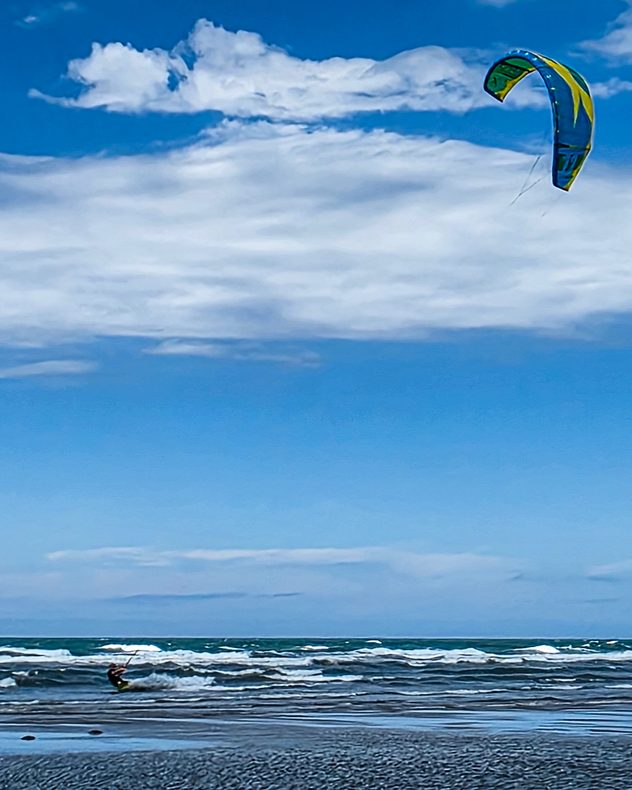 Kitesurfer riding choppy waves beneath layered white clouds and a deep blue sky, with a colourful kite soaring overhead above a wideswept beach and rough sea.