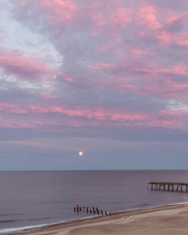 Full moon rising above a calm sea beneath pink and purple sunset clouds, with a sandy beach, wooden groynes, and a pier extending into the water on the right.
