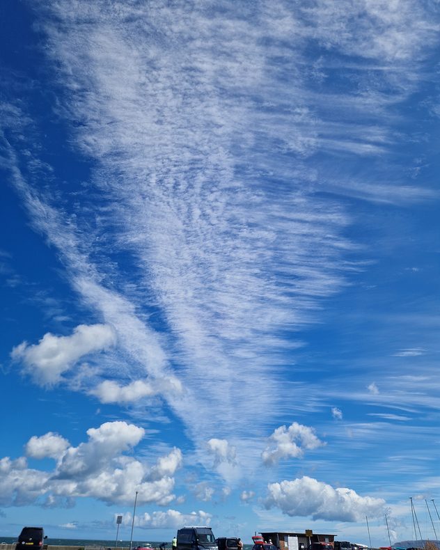 Blue sky filled with sweeping wispy cirrus streaks and rippled altocumulus cloud bands above scattered fair-weather cumulus clouds near a coastal car park and marina.