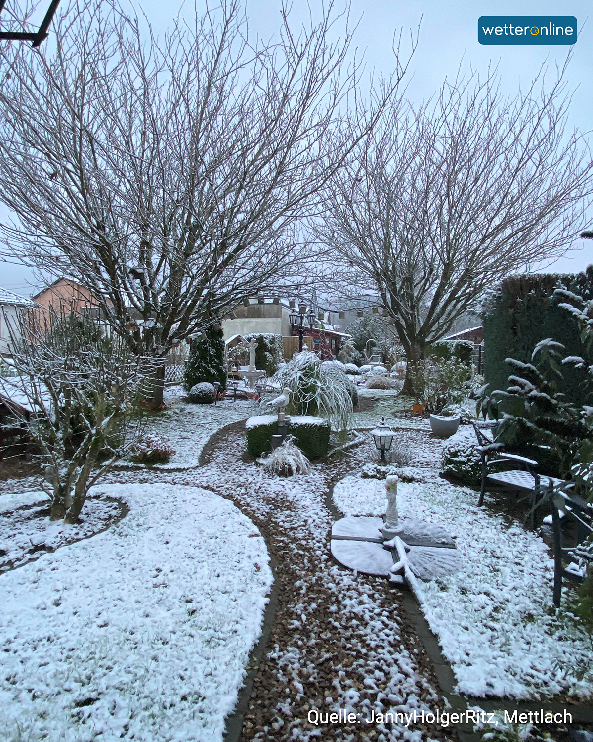 Verschneiter Garten mit Wegen, Sträuchern und kahlen Bäumen.