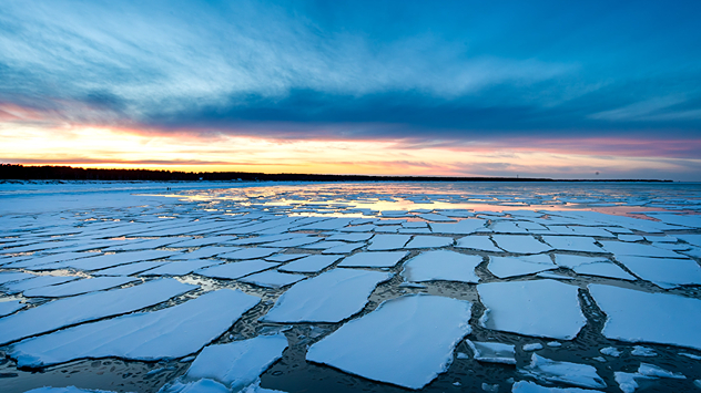 Aufgebrochene Eisschollen auf einem See bei Sonnenaufgang. Bunter Himmel über einer weiten Winterlandschaft.