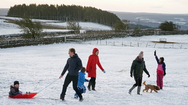Group of adults and children walking and playing on a snowy field, one child riding a red sled being pulled, others walking with a small dog, with winter hills and trees in the background.