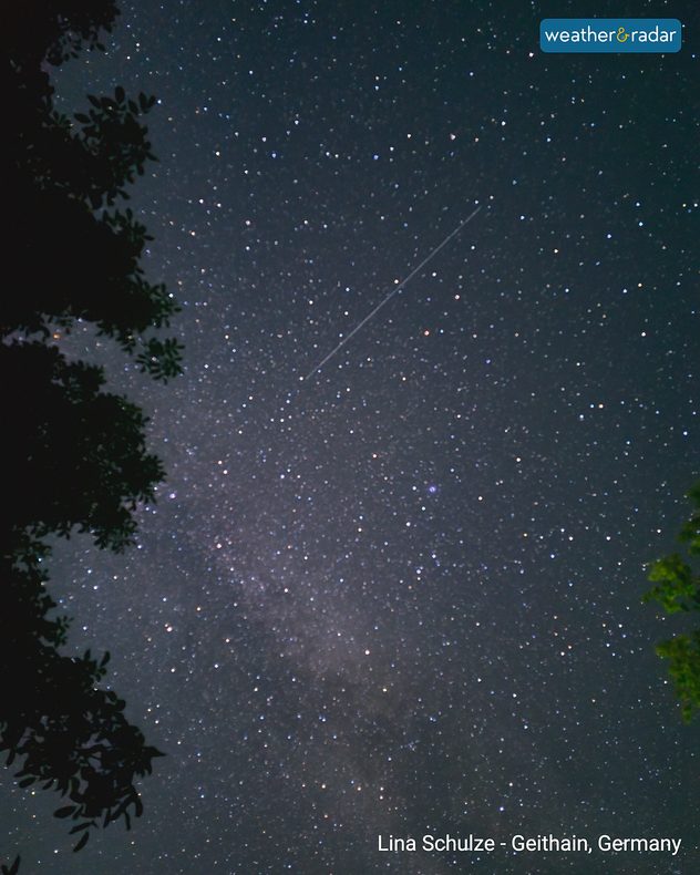 Bright meteor streak crossing a densely star-filled night sky above Geithain, Germany, with the Milky Way faintly visible and dark tree branches framing the view.
