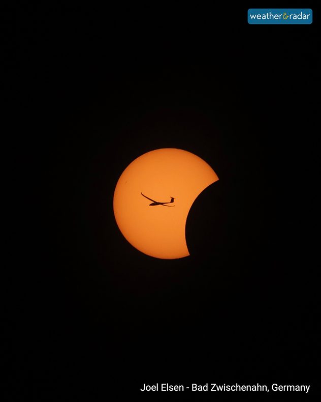 Orange Sun during a partial solar eclipse against a black sky, with the Moon obscuring its right side and a silhouetted aircraft crossing the solar disc.