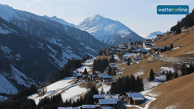 Alpines Dorf an einem Berghang mit teils schneebedeckten Wiesen, teils braunen Wiesen, dunklen Wäldern und einem hohen Gipfel im Hintergrund.