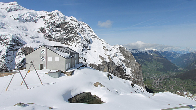 Eine Berghütte liegt in tief verschneiter Alpenlandschaft vor steilen Felswänden. Im Hintergrund reicht der Blick über ein grünes Tal und weitere Bergketten.