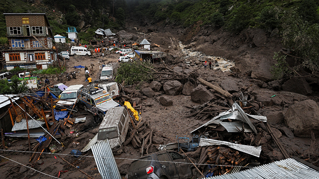Vue aérienne d'un village détruit après un glissement de terrain : voitures, maisons et débris ensevelis sous la boue, arbres et rochers dans le lit de la rivière.
