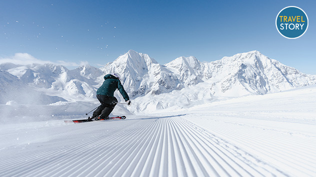 Ein Skifahrer fährt auf einer frisch präparierten Piste, im Hintergrund schneebedeckte Berge.