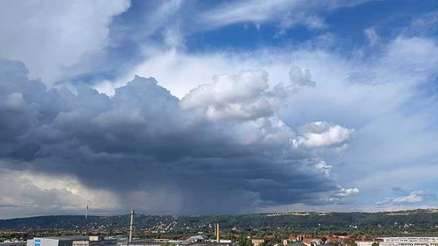 Dichte, dunkle Regenwolken ziehen über eine Stadt mit Industrie- und Wohngebäuden hinweg, im Hintergrund bewaldete Hügel.