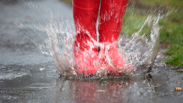 „Rote Gummistiefel springen in eine Pfütze, Wasser spritzt bei starkem Regen.