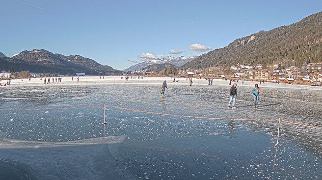 Das Webcambild zeigt den gefrorenen Weißensee mit einigen Eisläufern.