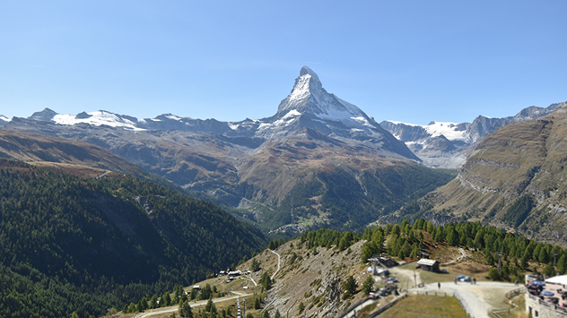 Das Bild zeigt das Matterhorn bei klarem, sonnigem Wetter. Im Vordergrund liegen Wälder, Wiesen und einige Berghütten.