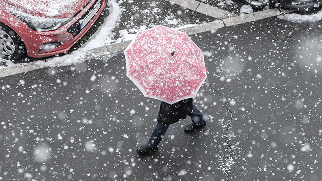 Das Bild zeigt eine Person mit rotem Regenschirm, die durch dichten Schneefall auf einer Straße geht. Am Straßenrand sind parkende Autos mit Schneeresten zu sehen.