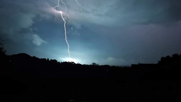 Bright lightning bolt striking beneath dark storm clouds over a silhouetted landscape at night, illuminating the sky with blue-white flashes.