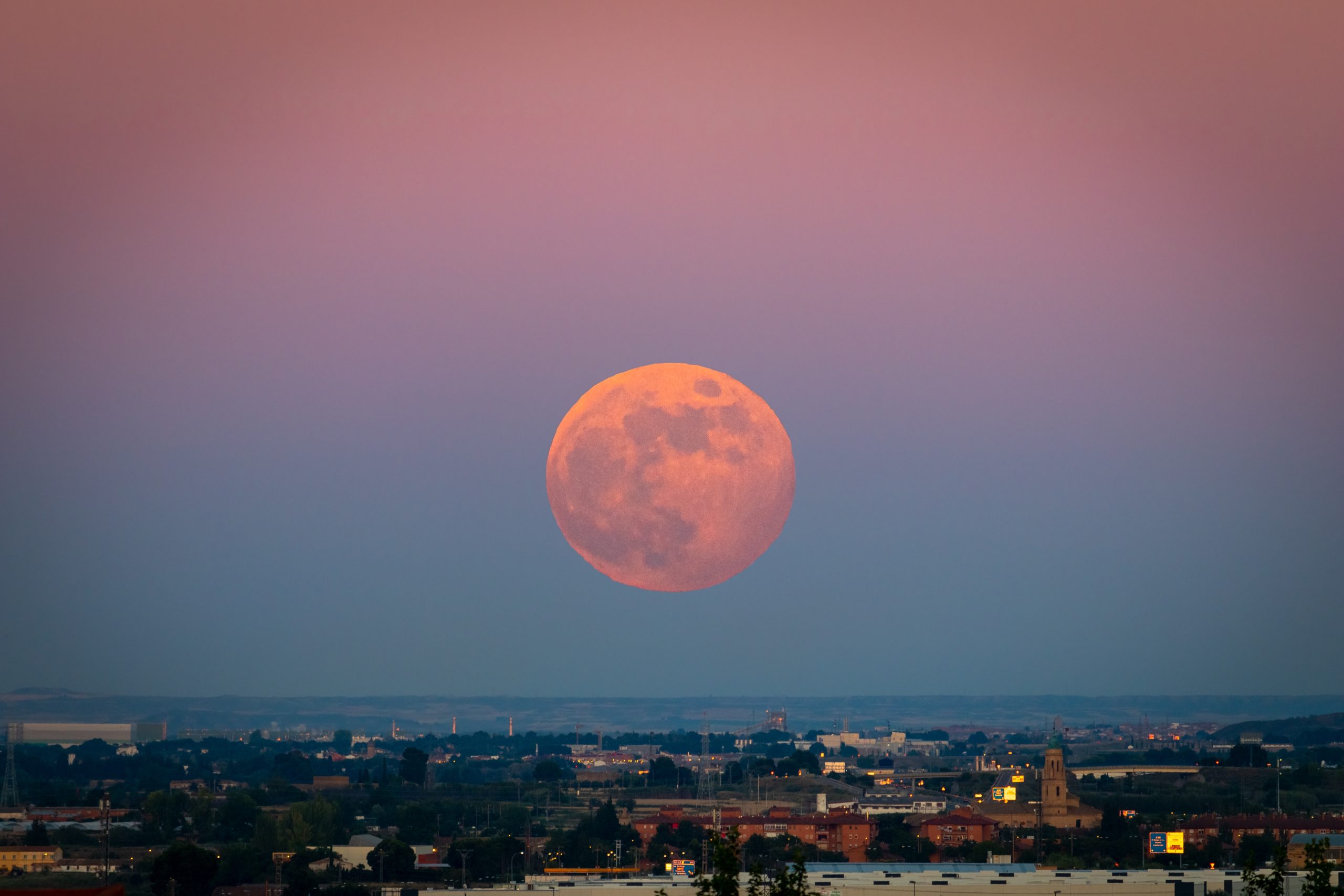 Full Strawberry Supermoon over city at sunset with purple and blue gradient sky. Urban nightscape photography with giant moon on the horizon, in June 2020 in Zaragoza, Spain.