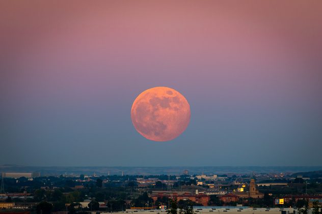 Full Strawberry Supermoon over city at sunset with purple and blue gradient sky. Urban nightscape photography with giant moon on the horizon, in June 2020 in Zaragoza, Spain.