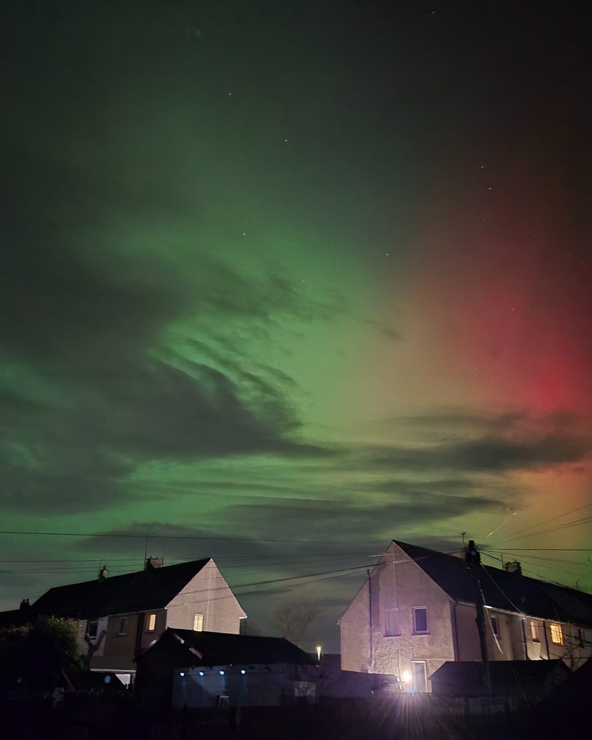 Night sky filled with green aurora curtains and a red glow to the right, with stars visible above silhouetted rooftops, power lines, and softly lit houses below.