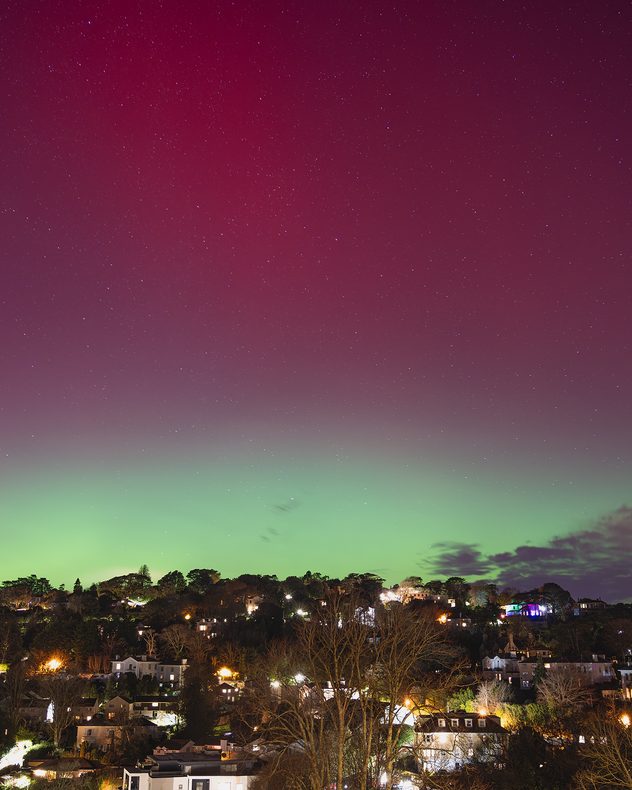 Wide night view of a town on a hillside with house lights below, beneath a star-filled sky glowing deep red above and bright green along the horizon from an aurora display.