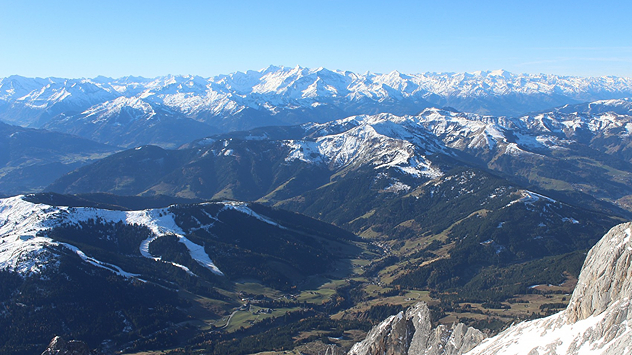 Das Bild zeigt eine weite Berglandschaft mit schneebedeckten Gipfeln und sonnigem Himmel. In den Tälern liegt nur wenig Schnee, und das Herbstlicht lässt die Landschaft klar erstrahlen.