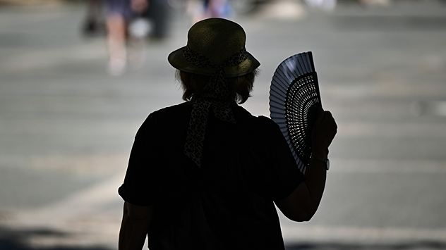 Une personne coiffée d'un chapeau se tient à l'ombre et s'éventaille avec un éventail.