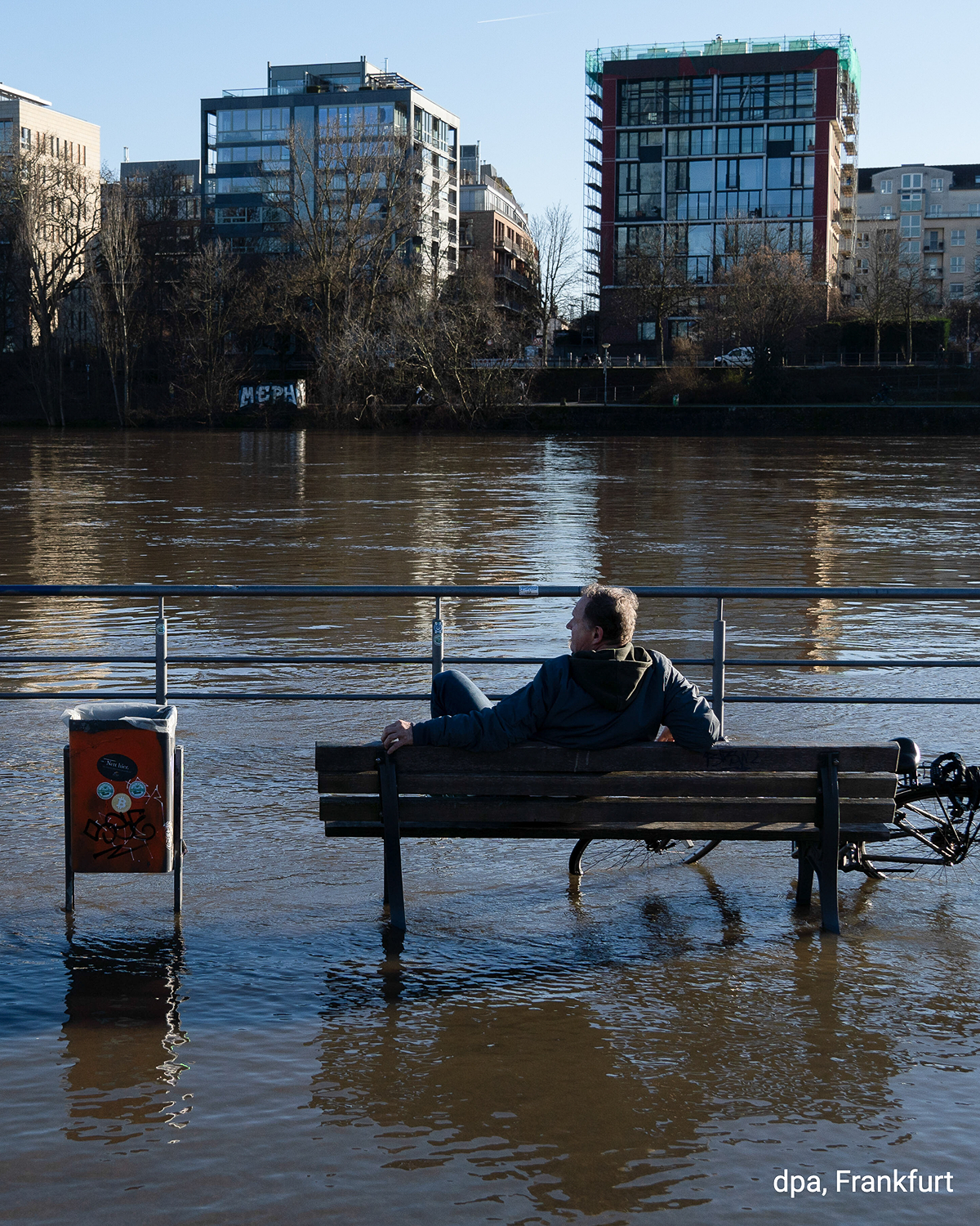 Mann sitzt auf einer teilweise überfluteten Bank am Mainufer in Frankfurt. Hochwasser bedeckt den Boden rund um die Bank.