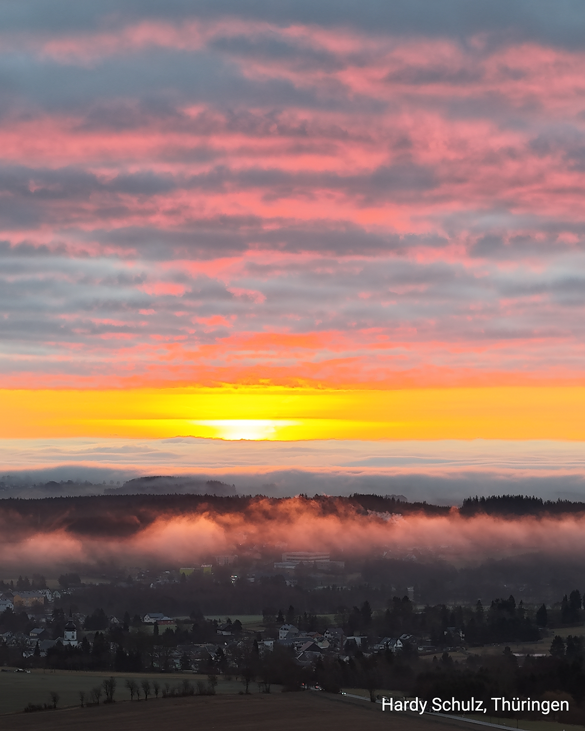 Sonnenaufgang mit rosa Wolken über einem nebeligen Ort in Thüringen. Goldenes Licht am Horizont über Hügeln und Wäldern.