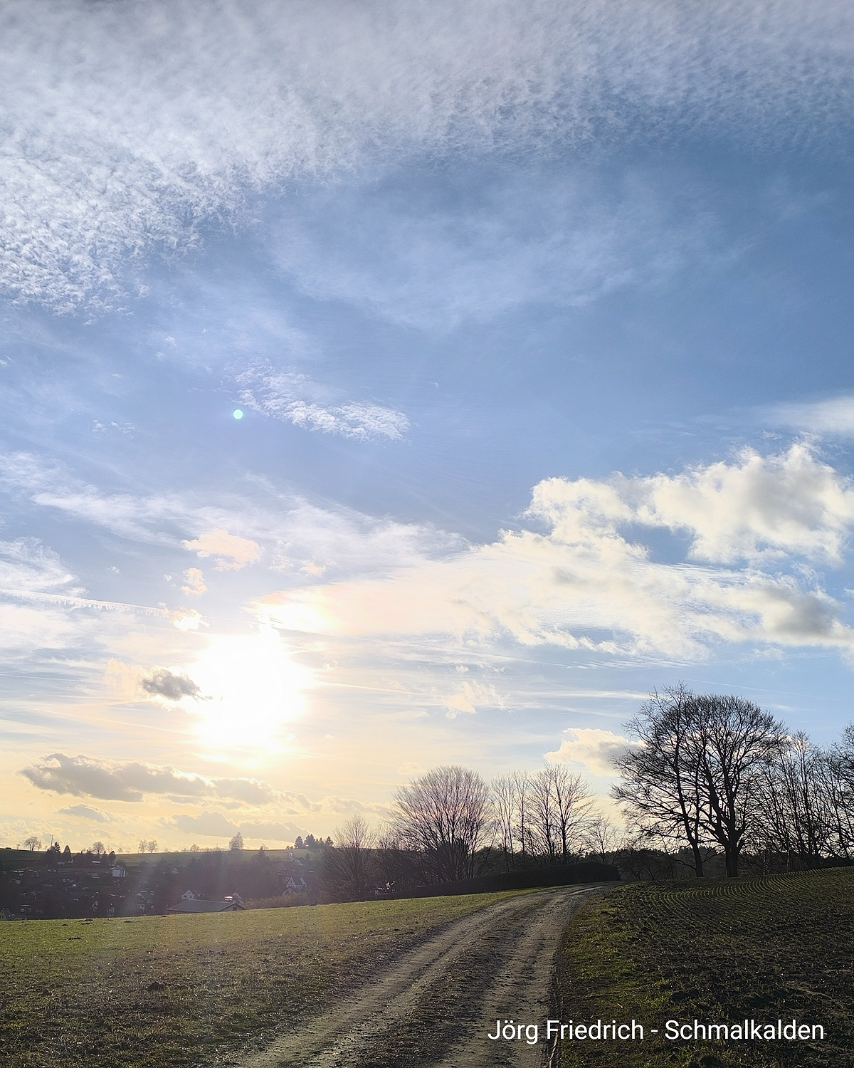 Feldweg in winterlicher Landschaft bei tief stehender Sonne. Kahle Bäume und wolkiger Himmel über einem Dorf am Horizont.
