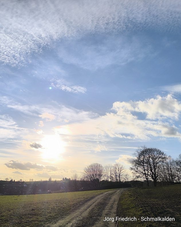 Feldweg in winterlicher Landschaft bei tief stehender Sonne. Kahle Bäume und wolkiger Himmel über einem Dorf am Horizont.