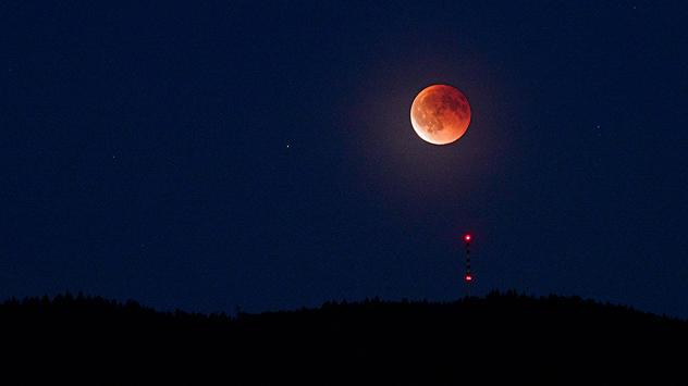 Roter Mond während einer Finsternis über der Hornisgrinde. Ein Funkturm mit rotem Licht steht silhouettenhaft im Vordergrund.