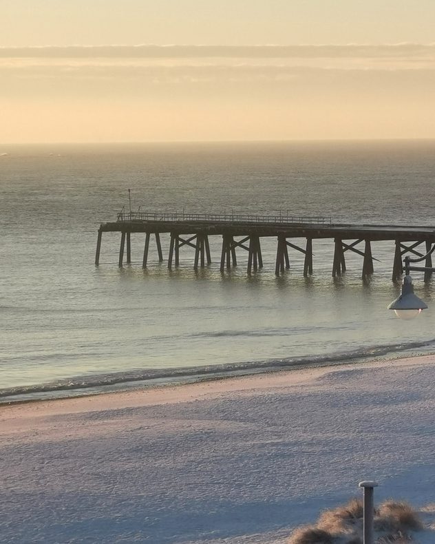 Wooden pier extending into a calm sea beside a snow-covered beach at sunrise, with gentle waves, pale golden light on the horizon, and soft pastel tones in the winter sky.